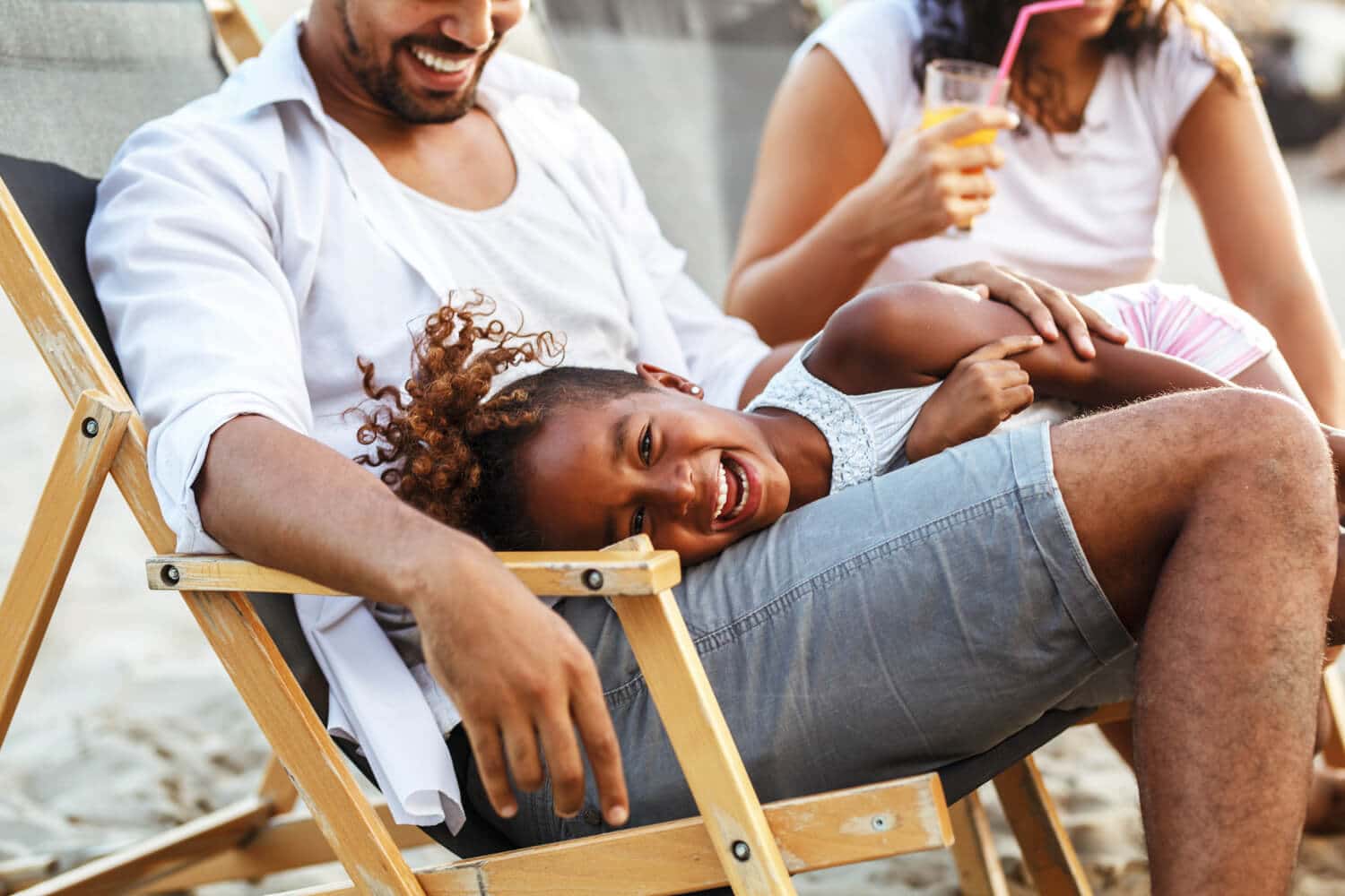 2 adults and a child sitting in chairs with a drink while relaxing on the beach.