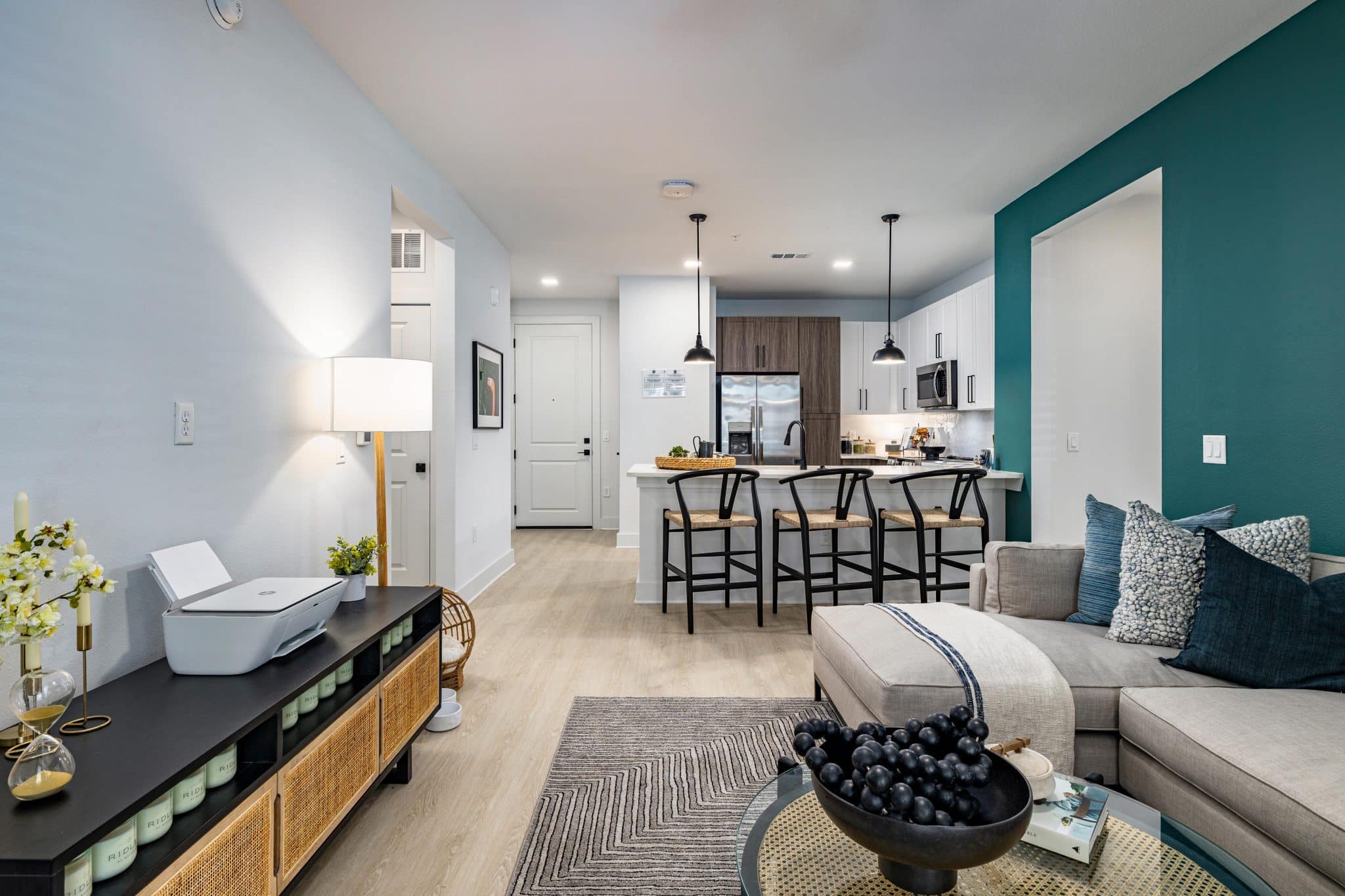 A living room with with hardwood-style flooring and open access to a kitchen with a breakfast bar near an entry door.