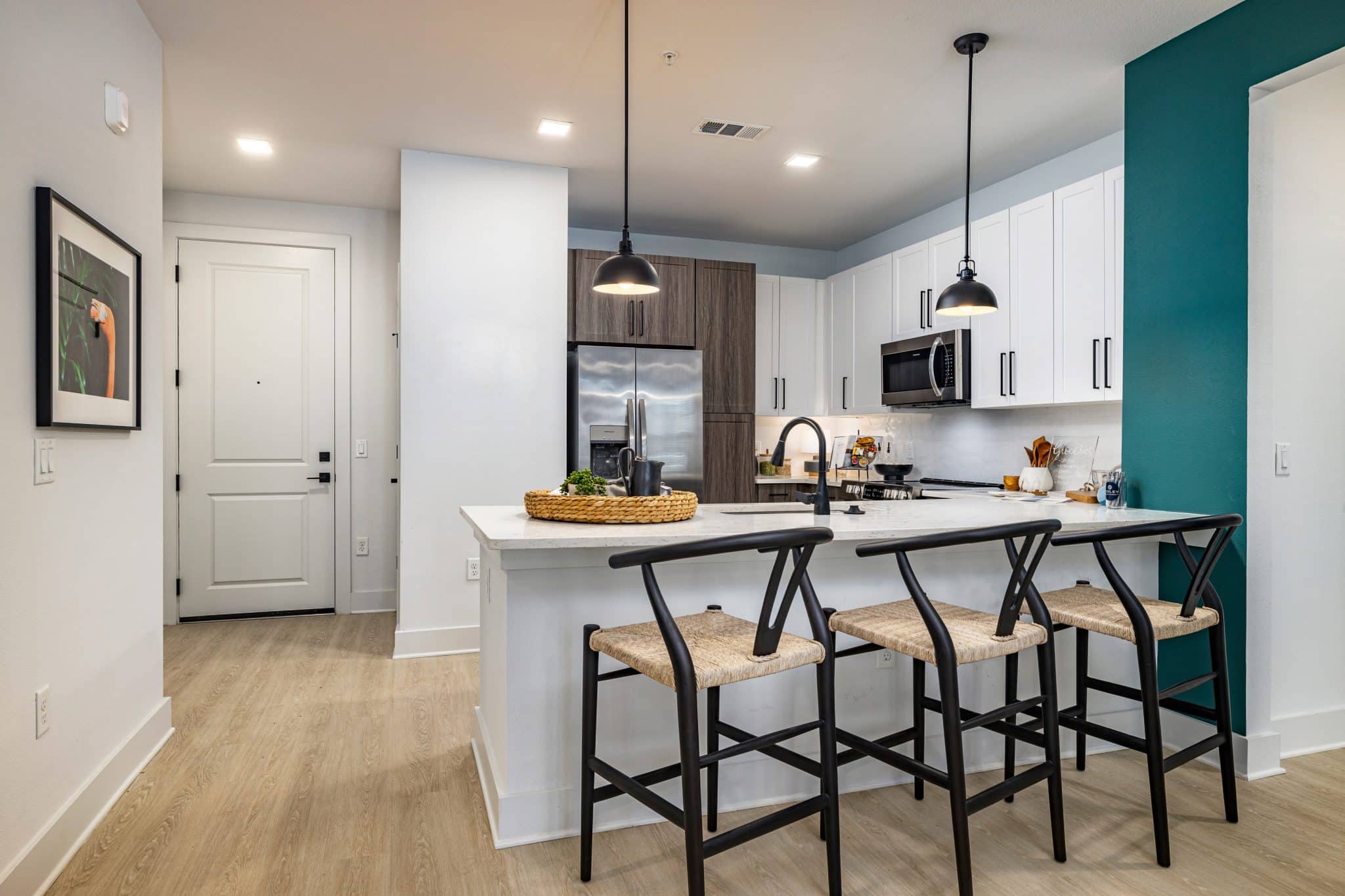 A kitchen with a breakfast bar, barstools, pendant lights, stainless steel appliances, and cabinets near an entrance door.
