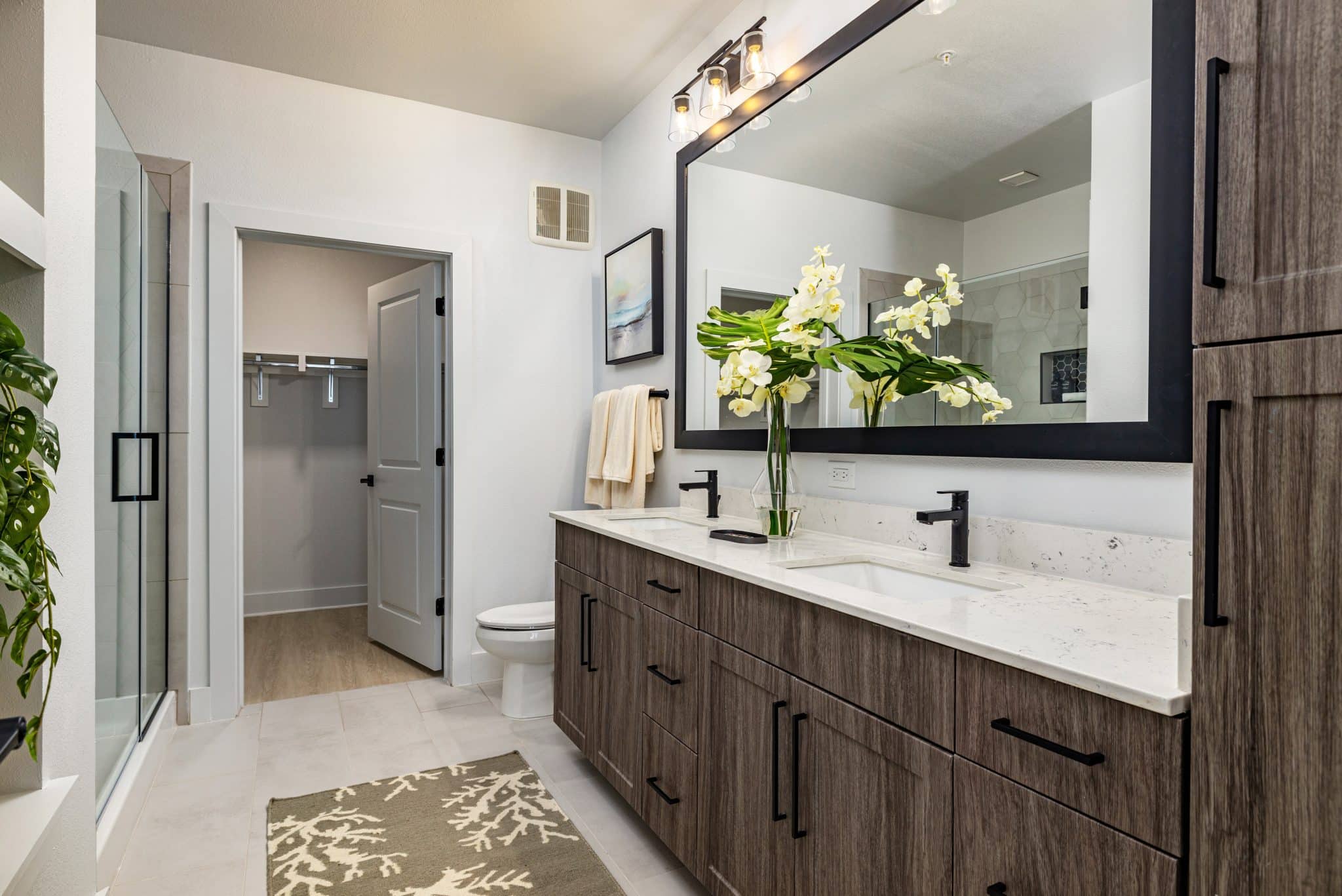 A bathroom with a shower, double vanity with quartz countertops, large mirror, toilet, walk-in closet, and tile floors.