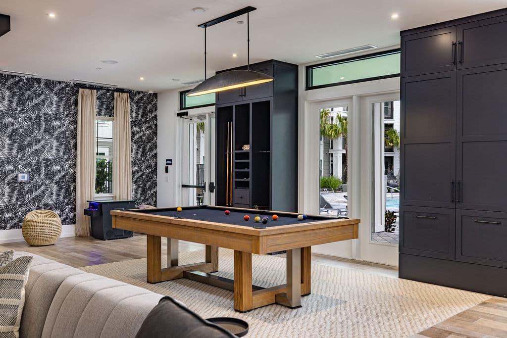 A billiards room with a pool table, a window, and doors leading to the outdoor pool area at Ridley at Waterset.