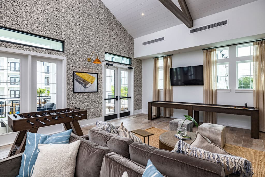 A community clubhouse with comfortable sofas, a foosball table, and a shuffleboard table near doors and windows.