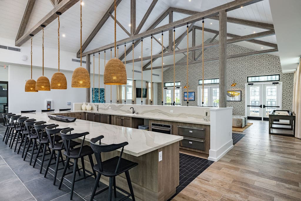 A large community kitchen with a bar, barstools, and pendant lights near a lounge with a shuffleboard at Ridley at Waterset.