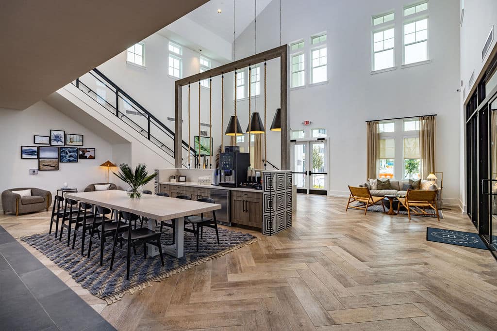 The interior of a 2-story clubhouse with several windows, seating areas, a coffee station, a staircase, and exit doors.