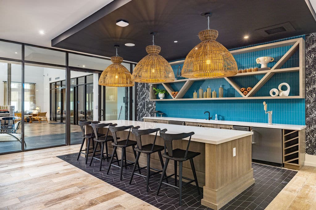 A clubhouse kitchen with a bar, barstools, rattan hanging lights, and a glass wall near a seating lounge.