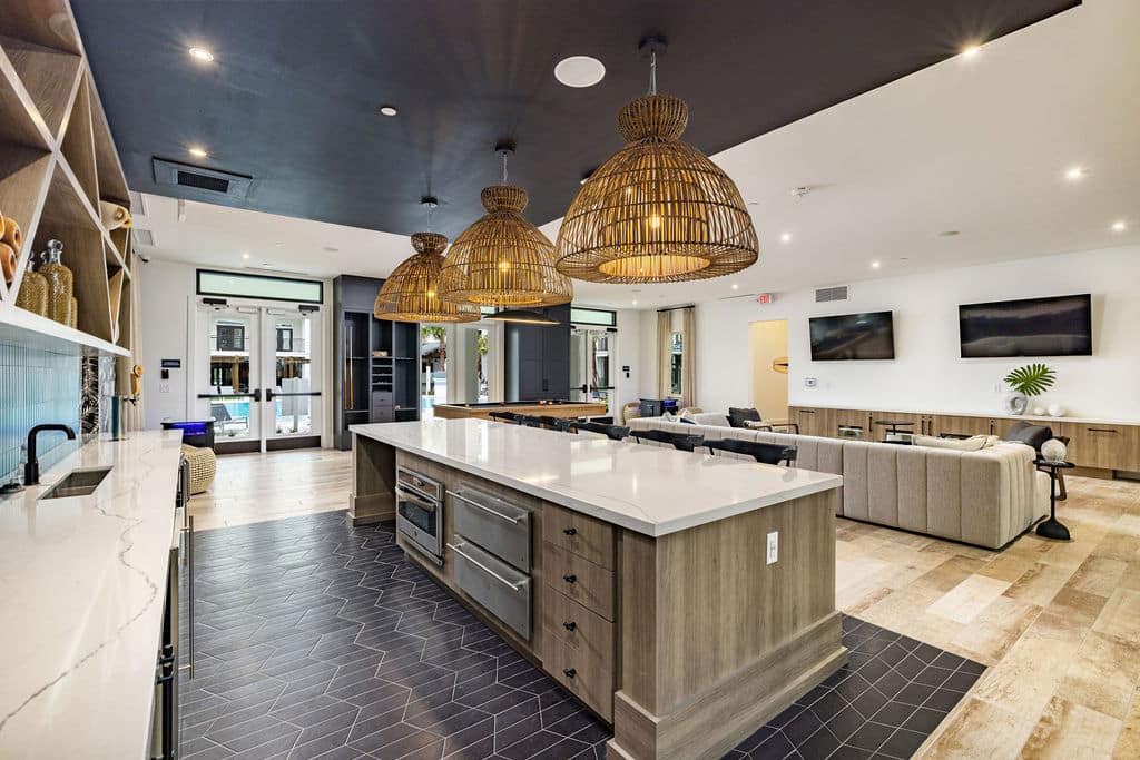 A community kitchen with quartz countertops and an island with rattan pendent lights near a seating area with TVs.