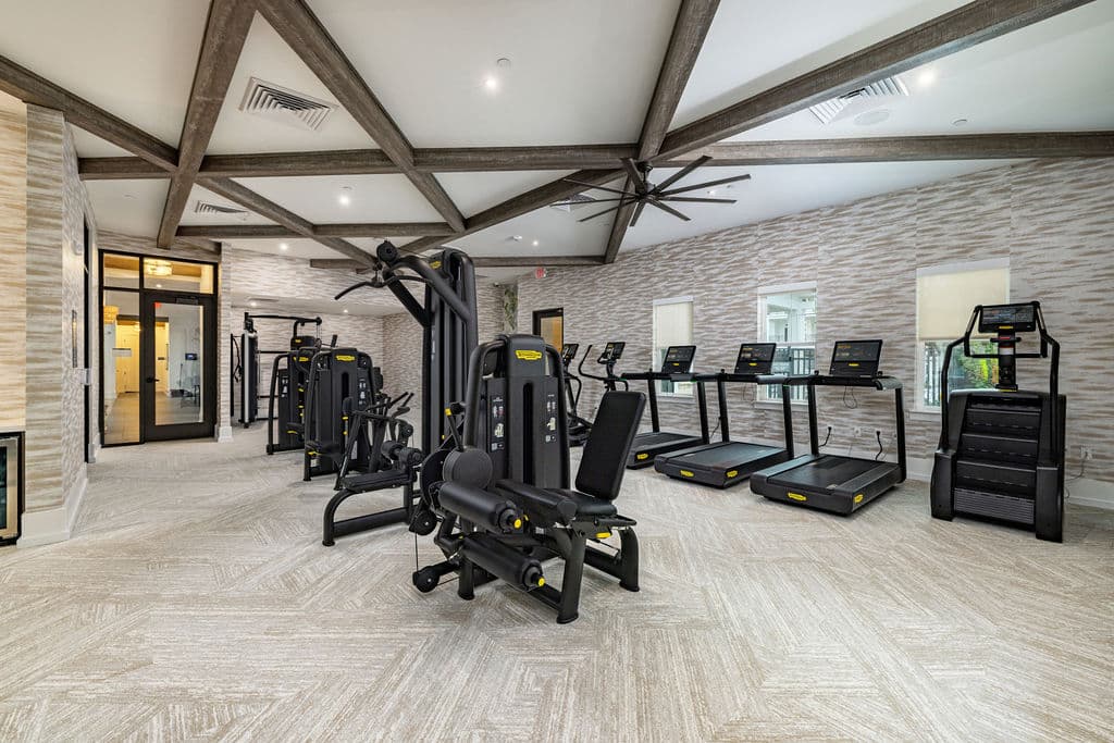 A modern fitness center with cardio and weight equipment, ceiling fans, windows, and wooden beams on the ceiling.