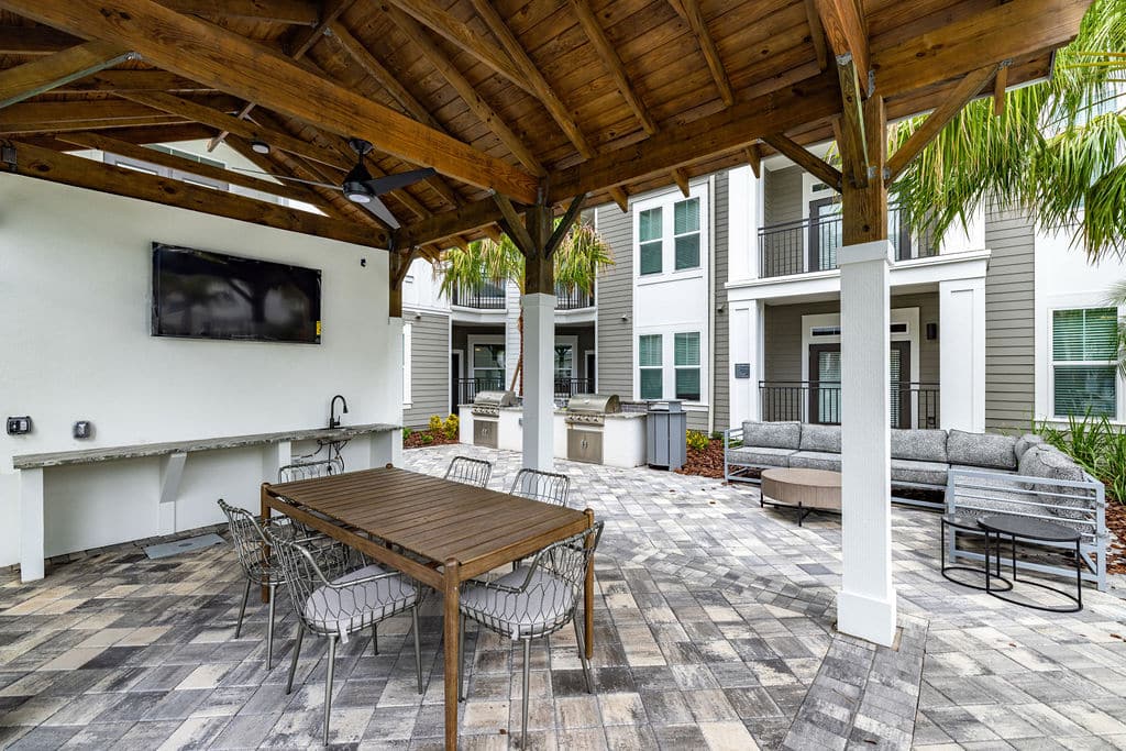 Covered outdoor entertainment area with a table, chairs, and a TV near a grilling station and pool.