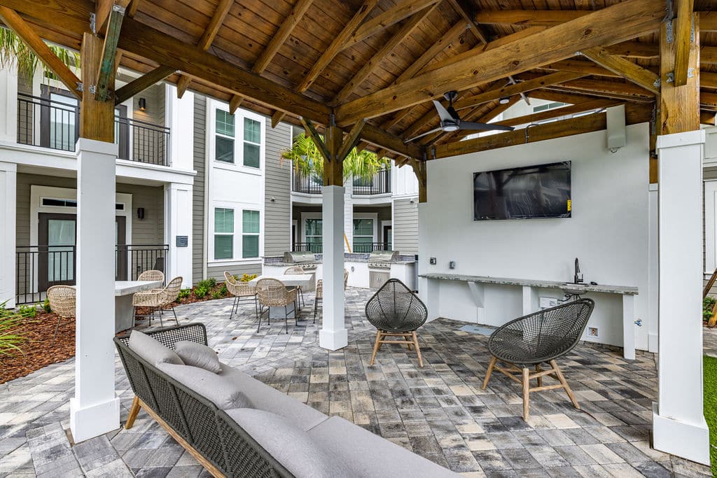 A covered entertainment lounge with a sofa, chairs, and tables near a grilling station at Ridley at Waterset.