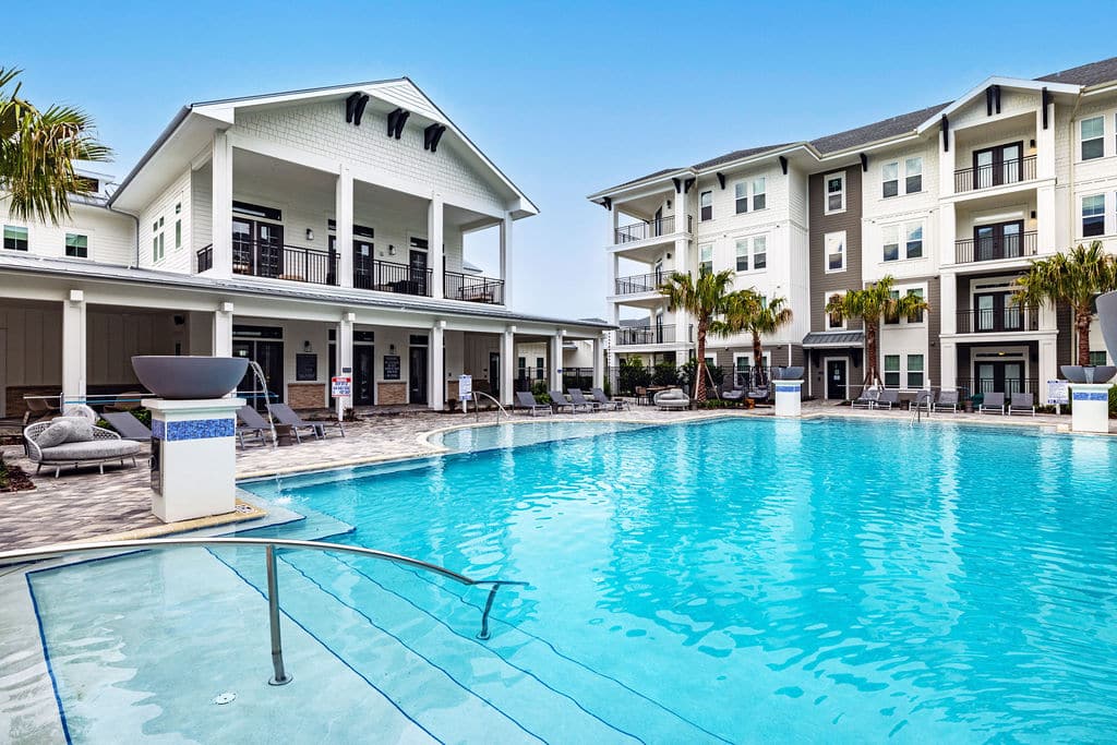 A resort-style pool with fountains and lounge chairs near a community clubhouse and apartments at Ridley at Waterset.