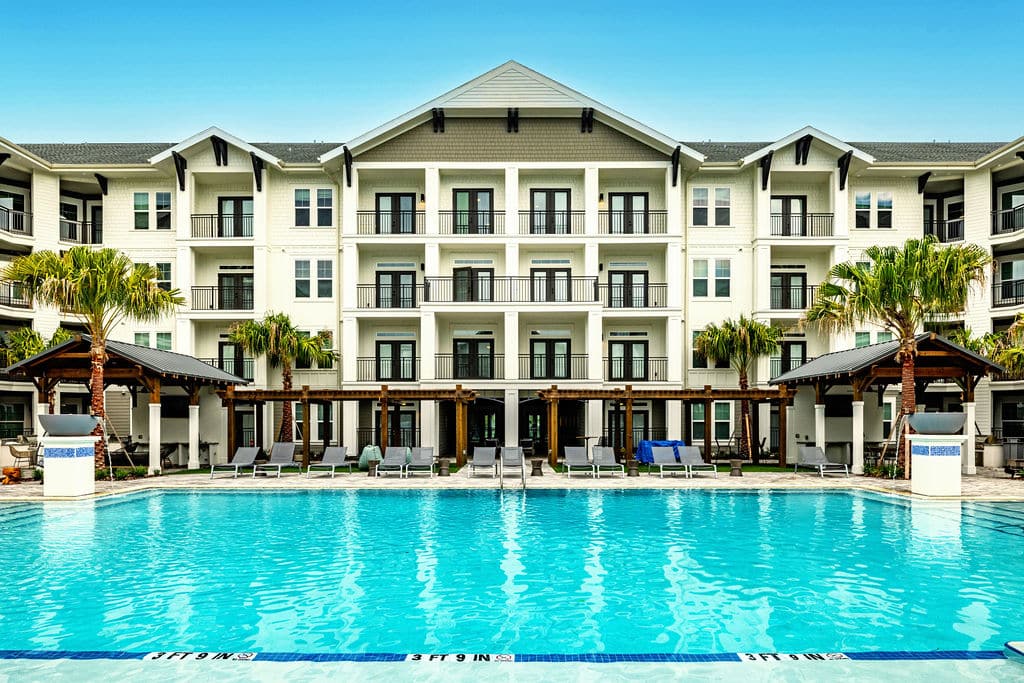 A resort-style pool with fountains, lounge chairs, and cabanas, surrounded by apartments with balconies.