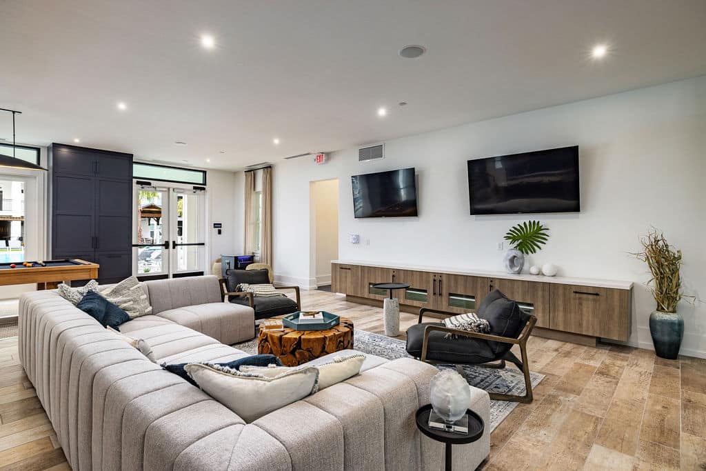 A modern community lounge with a large sectional, chairs, and TVs near a shuffleboard and doors to a pool.